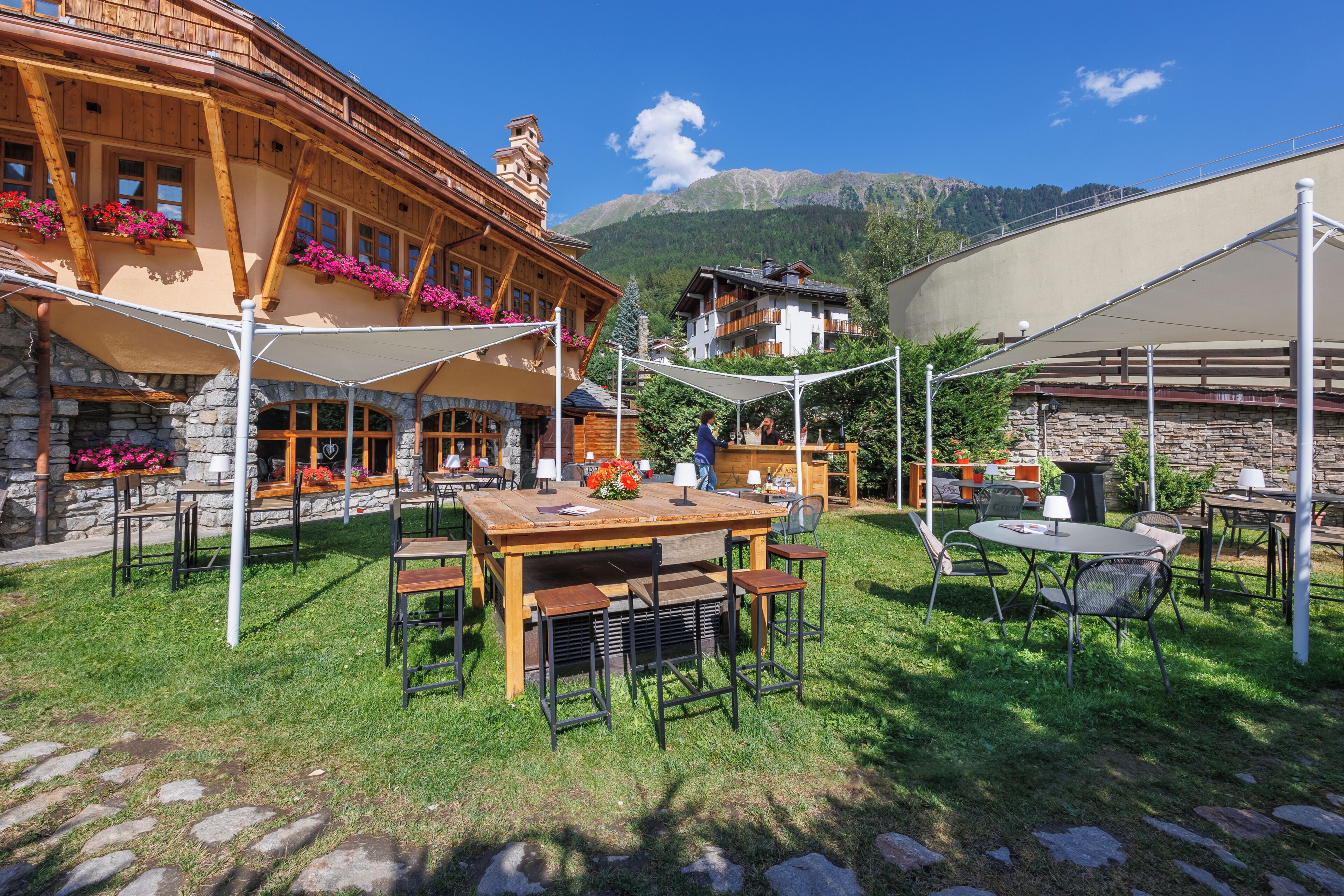 Ein Essbereich im Freien mit Tischen und Stühlen, ein Steinhaus mit Holzbalken und eine Bergkulisse im Hintergrund.