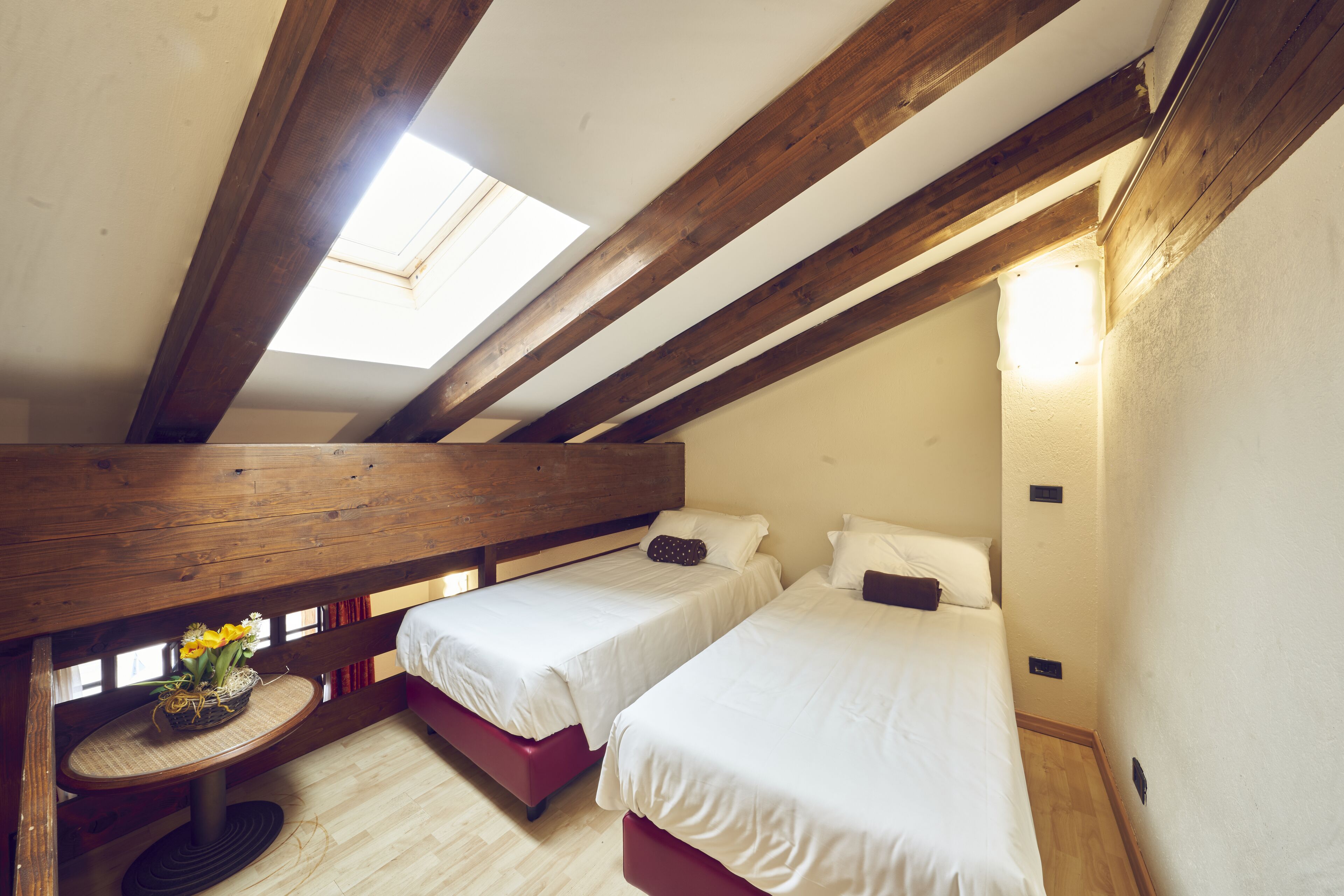 Une chambre avec deux lits, des poutres apparentes, une lucarne et une petite table sur laquelle est posé un vase.