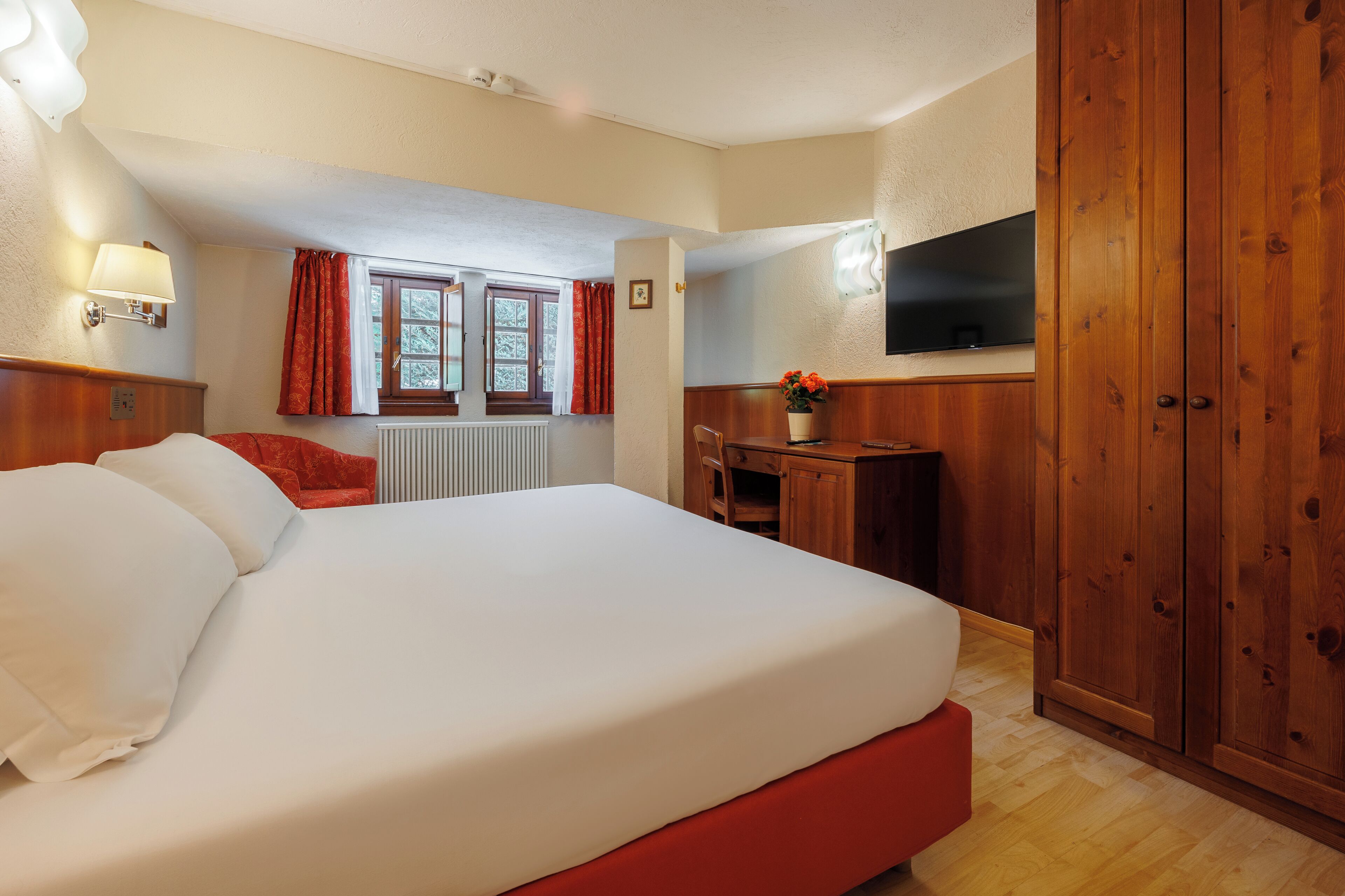 A hotel room with a large bed, a desk, a chair, a TV, and wooden wardrobe.