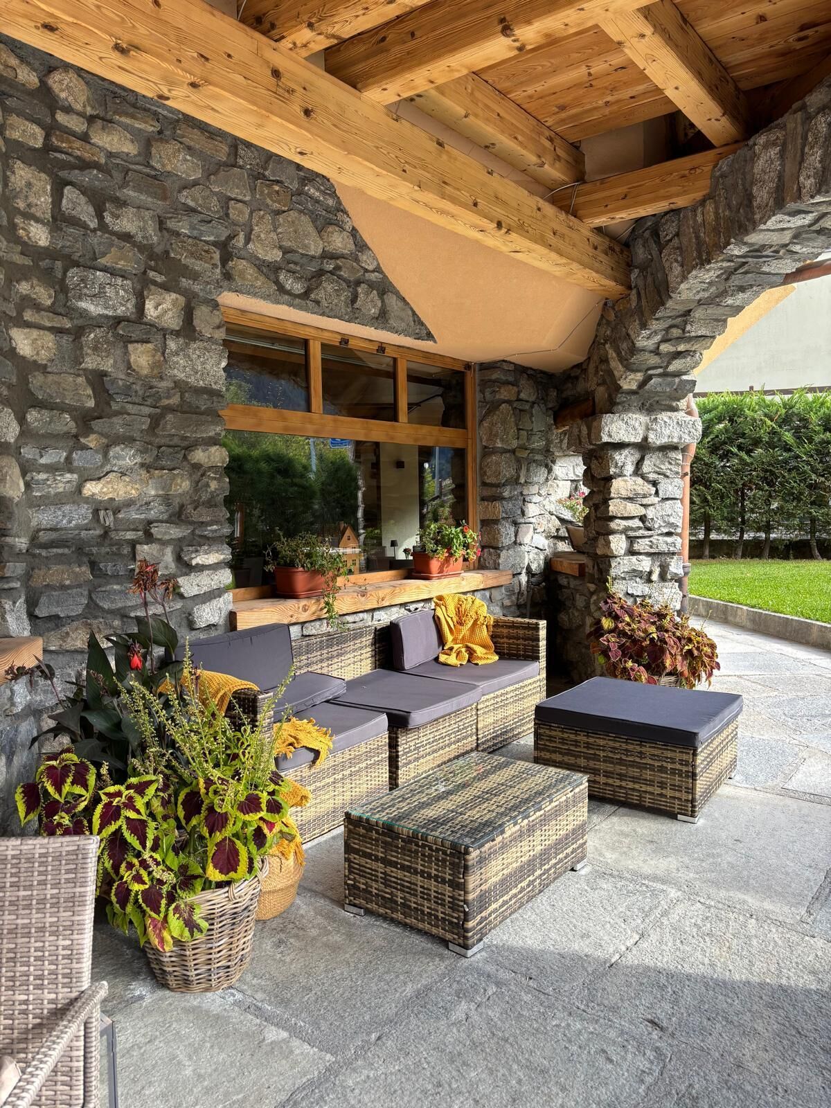 Une façade en pierre, un plafond en bois, des sièges d’extérieur avec des coussins et des plantes en pot.