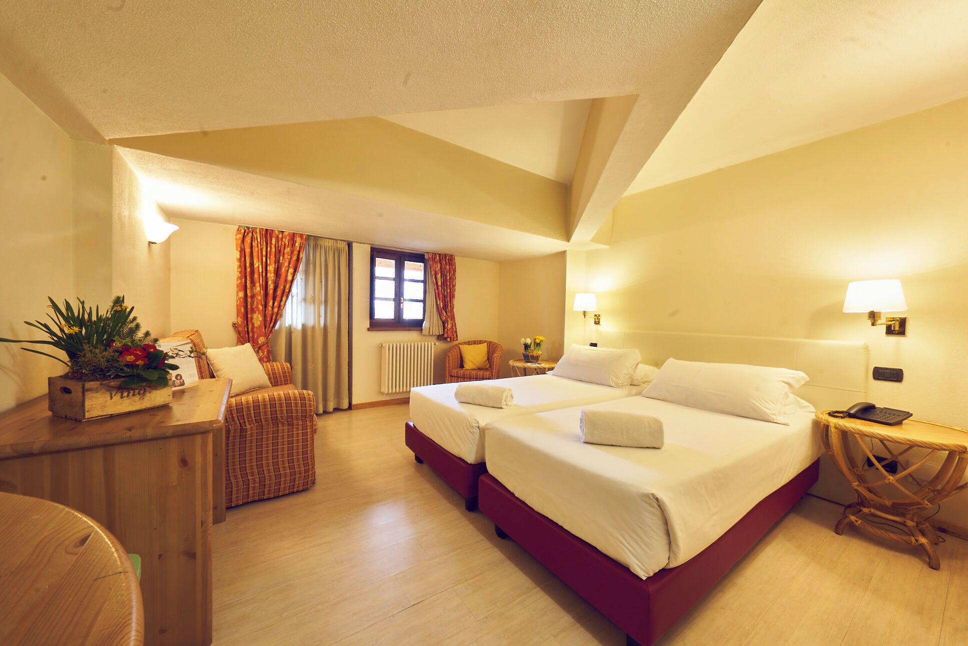 A hotel room with two beds, a wooden desk, a chair, a small table with a lamp, and a window with curtains.