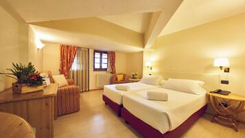 A hotel room with two beds, a wooden desk, a chair, a small table with a lamp, and a window with curtains.