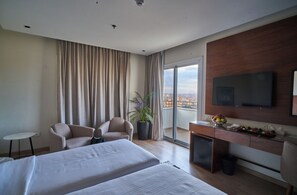A hotel room with two beds, a desk with a TV, a chair, a plant, and a view of the city.