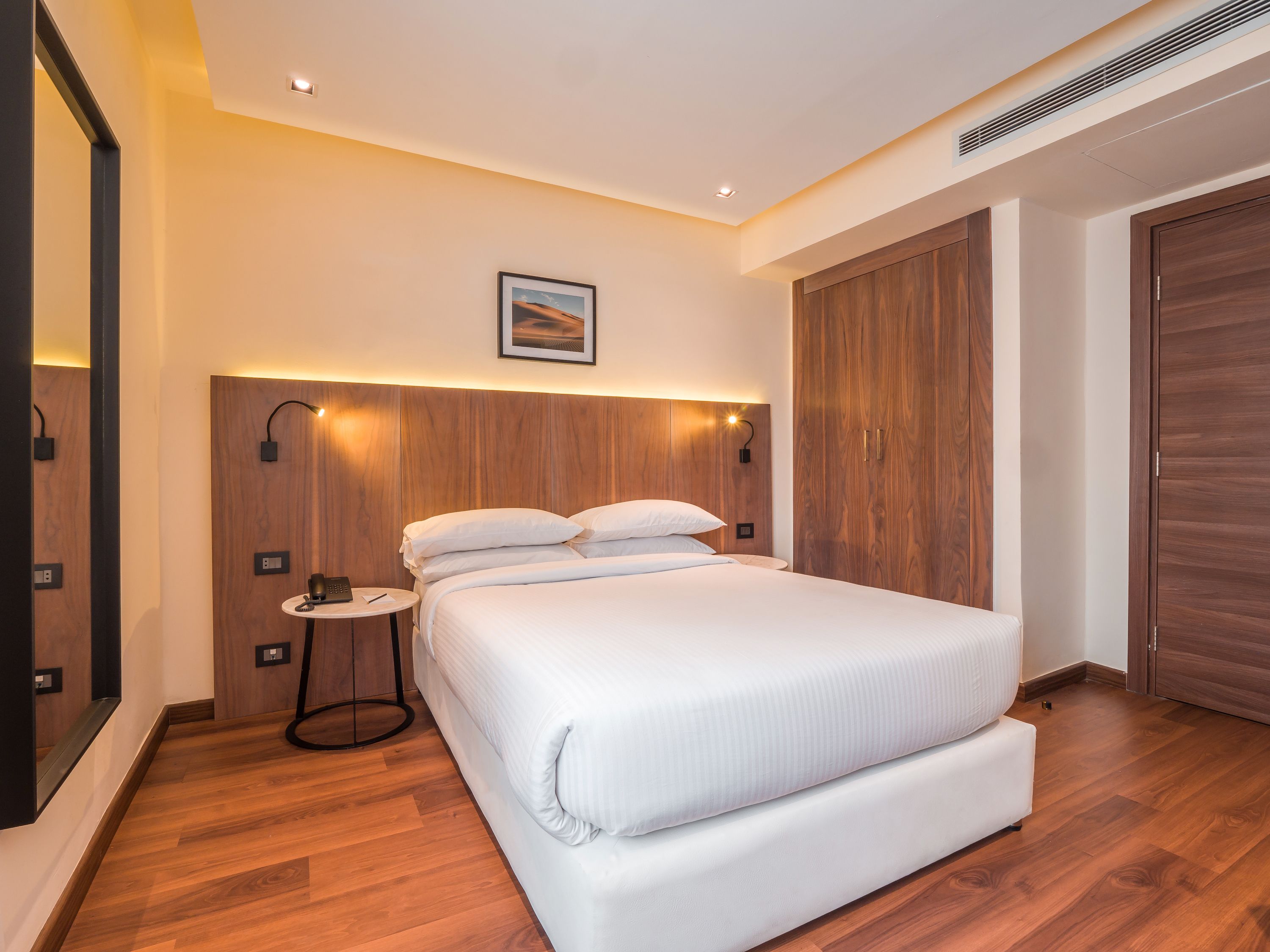 A modern hotel room with a large bed, wooden headboard, bedside tables, and a wardrobe.