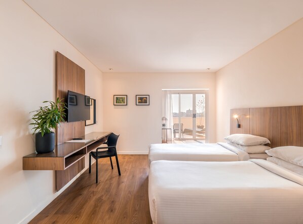 Habitación de hotel moderna con un escritorio de madera, televisión, dos camas, balcón con vistas y una planta en maceta.