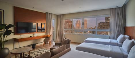 Habitación de hotel con televisión, escritorio, dos camas, sofá y vista panorámica de la ciudad.