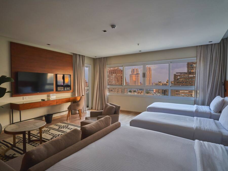 A hotel room with a TV, a desk, two beds, a sofa, and a view of the cityscape.