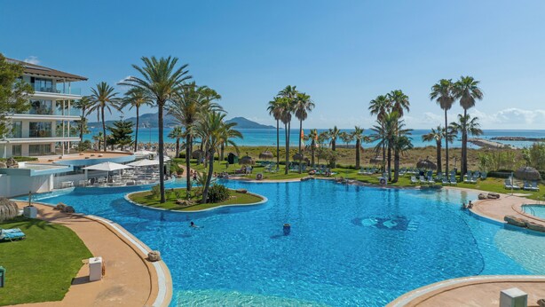 A large swimming pool with multiple sections, surrounded by palm trees and lounge chairs, with a view of the ocean and mountains in the background. - Playa Esperanza Resort (Muro)