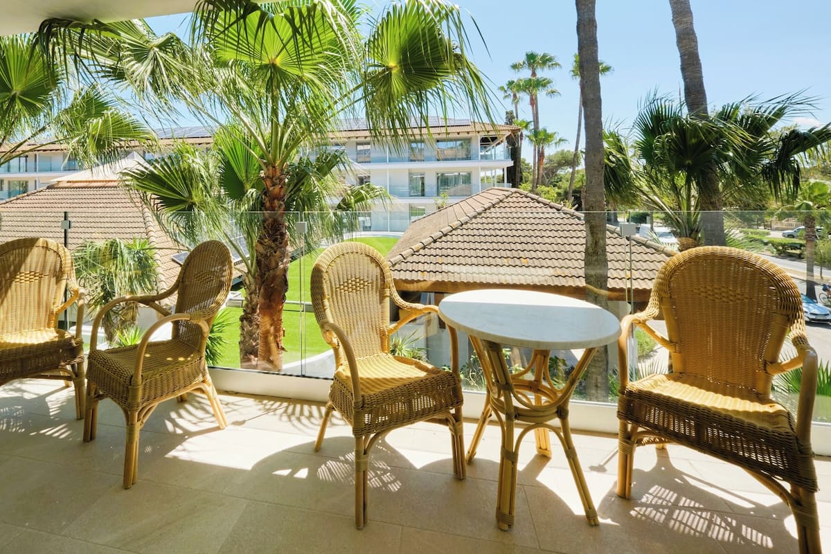 A balcony with wicker furniture, palm trees, and a view of a building with a tiled roof.