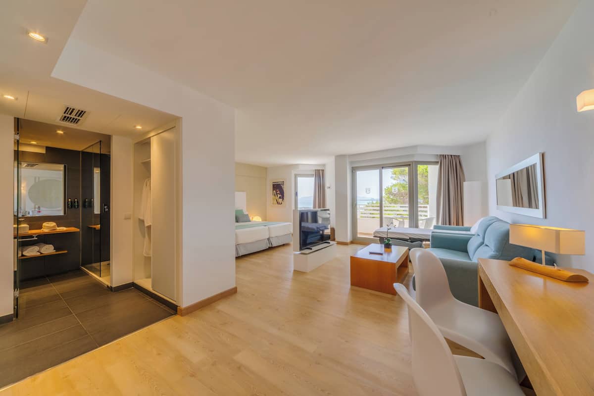 A modern hotel room with a wooden floor, a sofa, a TV, a dining table, and a large window with curtains.