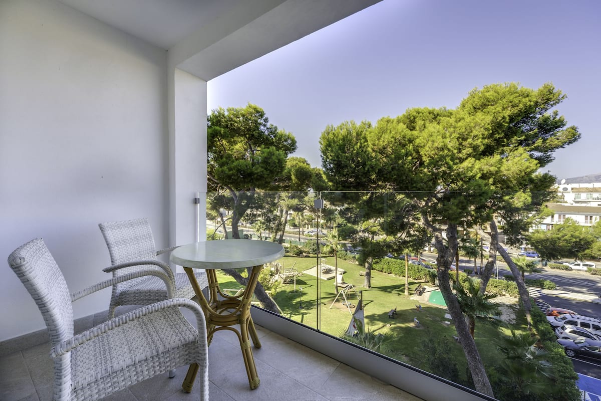 A balcony with wicker chairs and a glass table, overlooking a green landscape with trees and a parking area.