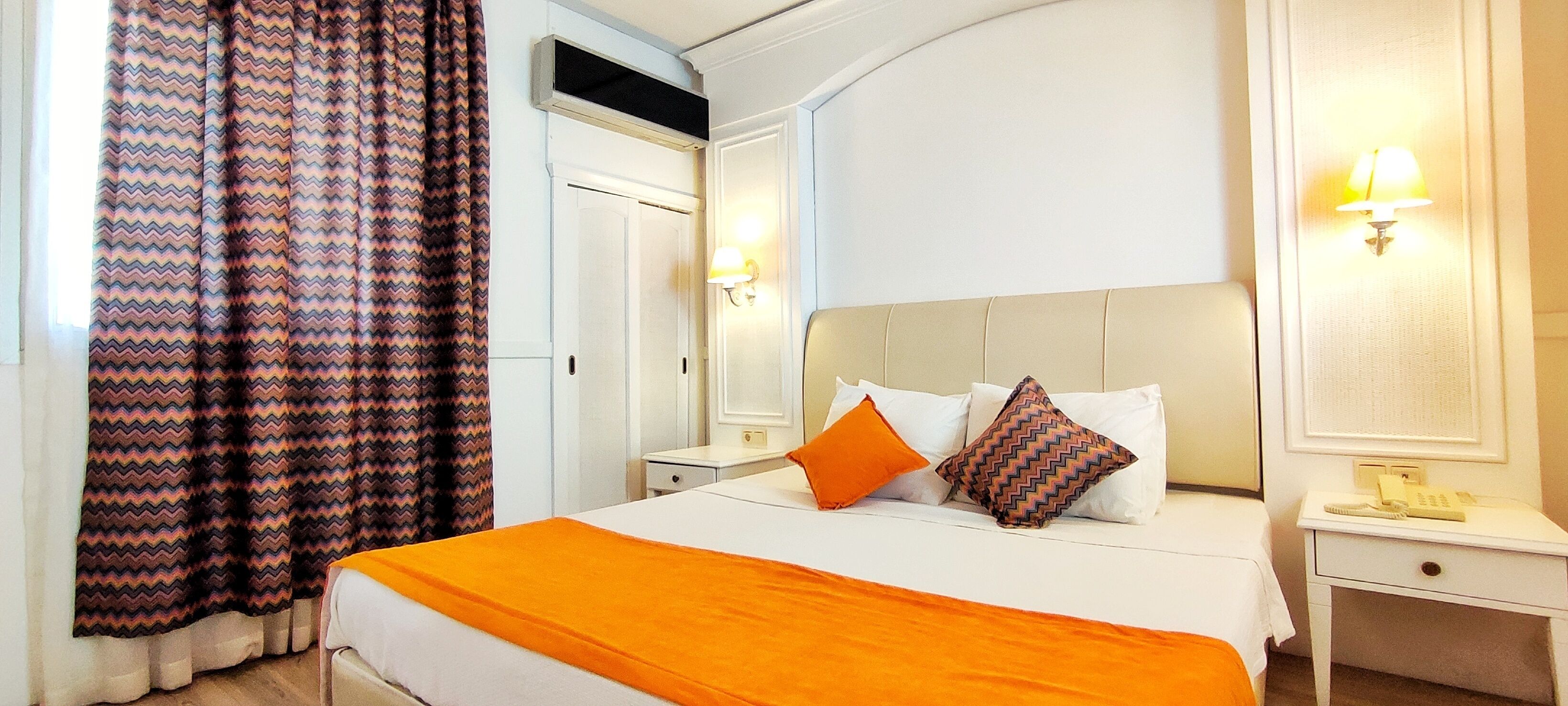Una habitación de hotel con una cama grande, dos mesitas de noche, un tocador, un espejo y una ventana con cortinas.