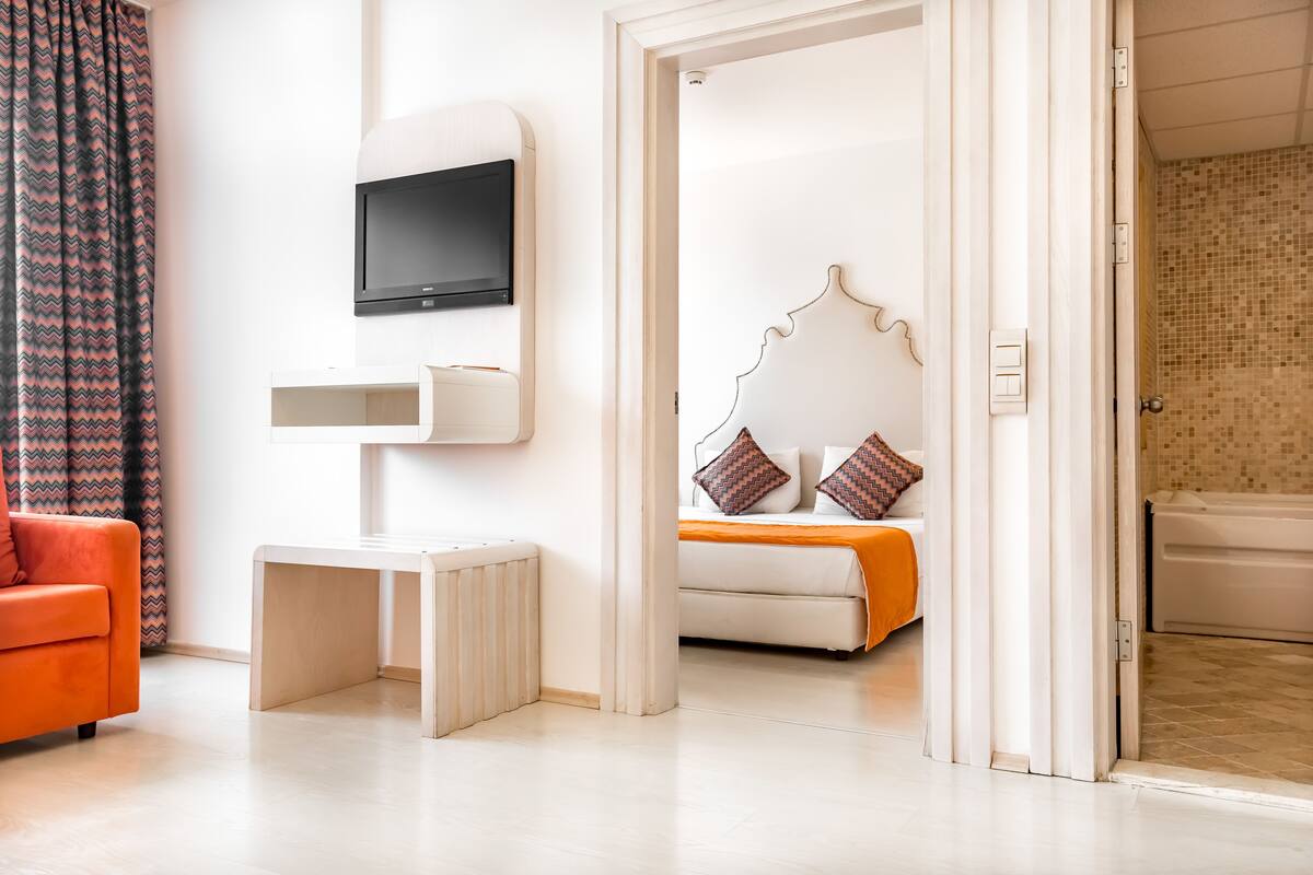 A modern hotel room with a bed, a television, a small table, and a bathroom visible through an open door.