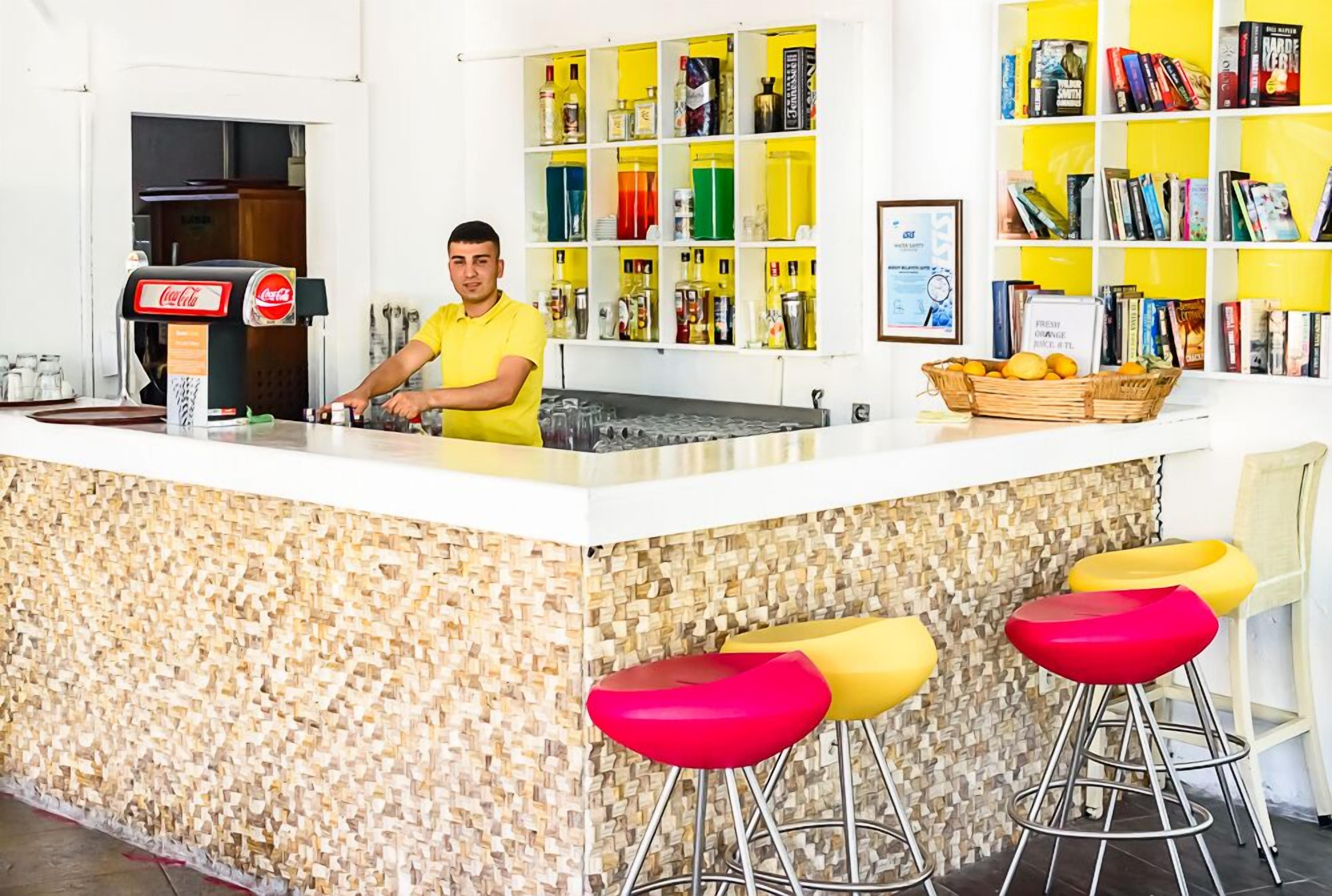 A bar area with a counter, a Coca-Cola dispenser, a variety of bottles, a bookshelf with books, and colorful stools.