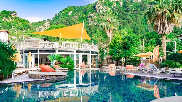 Zona de piscina con sillones de descanso y una cabaña, rodeada de exuberante vegetación y un telón de fondo montañoso.