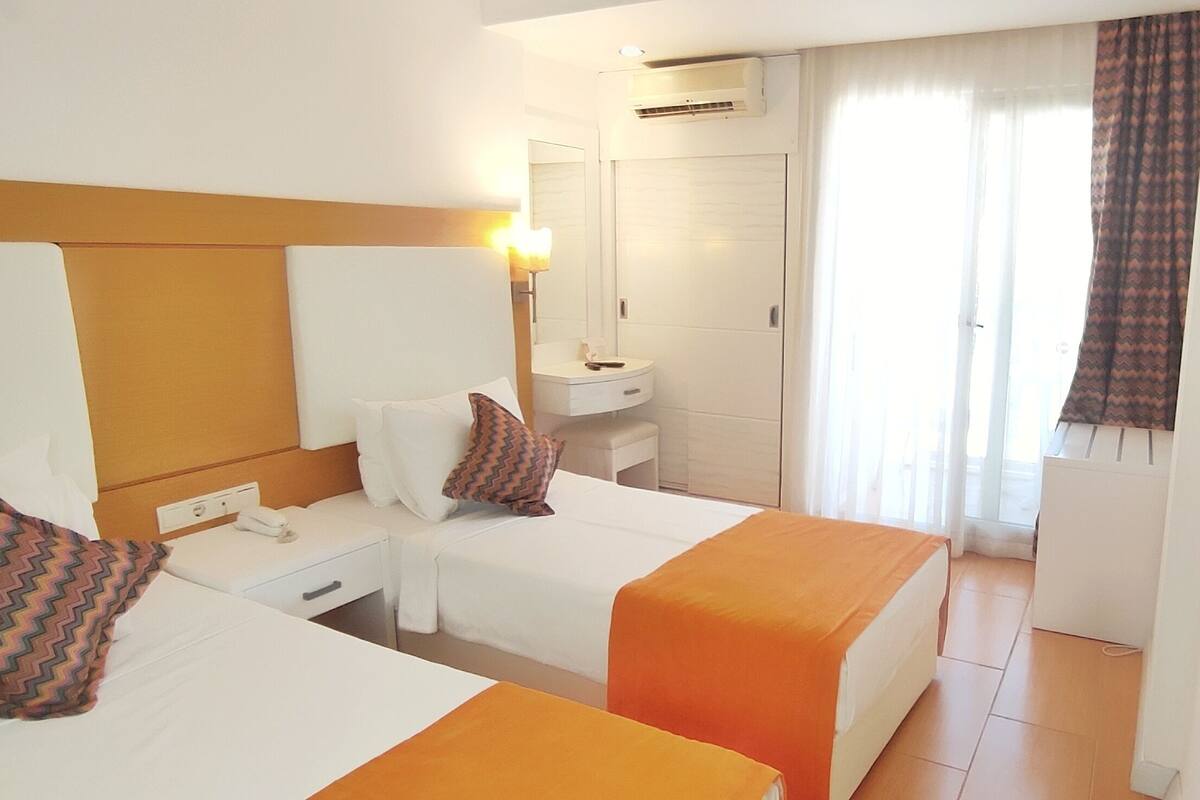 A hotel room with two beds, a bedside table, a mirror, a small bathroom, and a window with curtains.