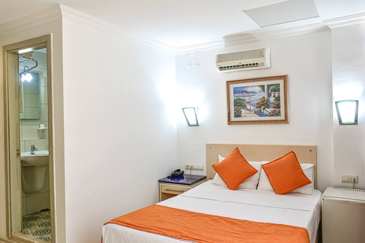 A hotel room with a bed, a nightstand, a desk, an air conditioner, a wall-mounted picture, and a bathroom visible through an open door.