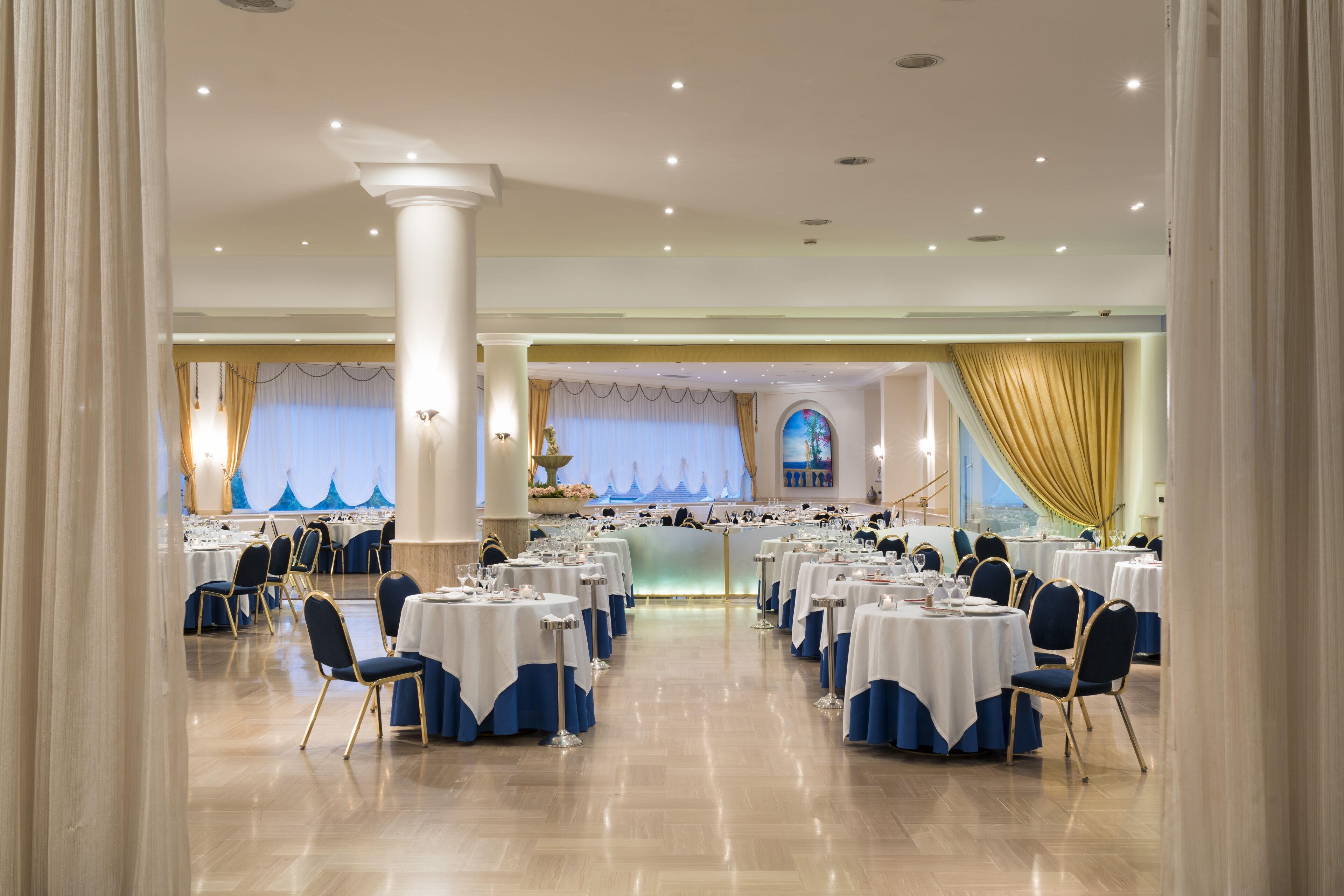 A formal dining room with round tables, blue and white tablecloths, and gold chairs.