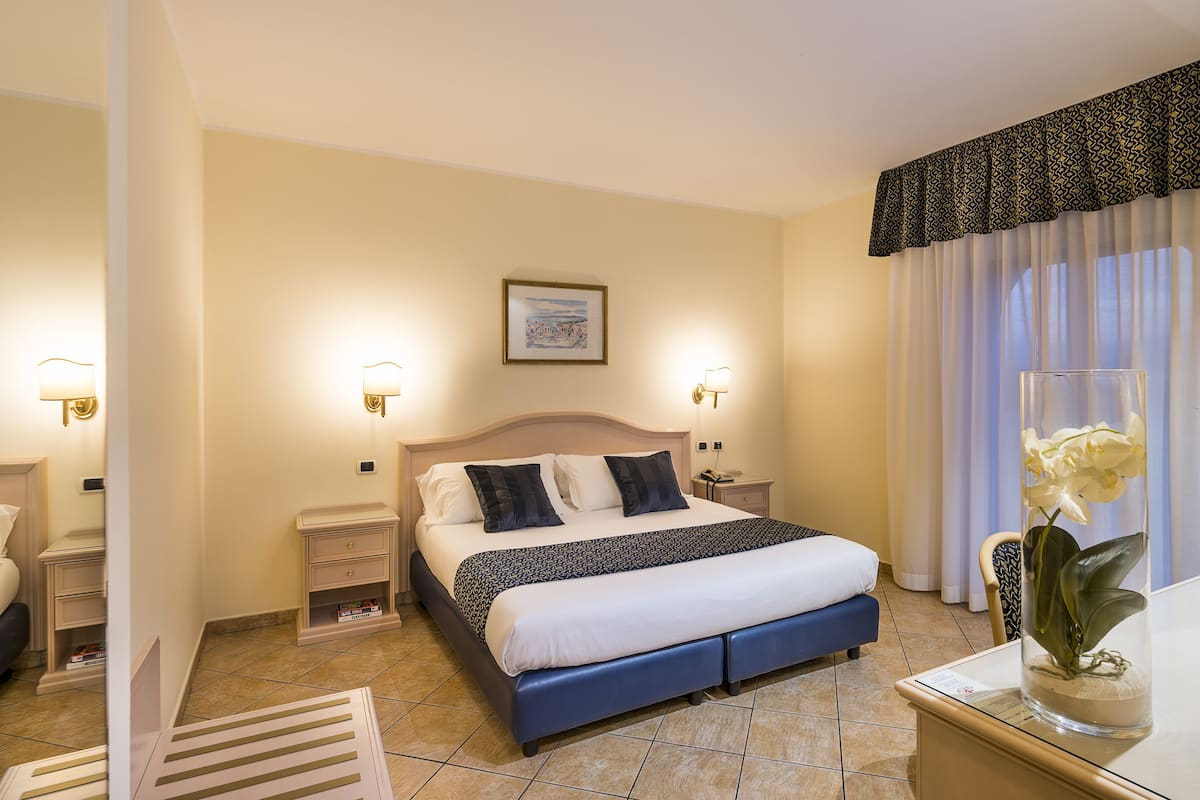 Ein Hotelzimmer mit einem großen Bett, Nachttischen, einem Beistelltisch, einem Sessel und einer Vase mit Blumen.