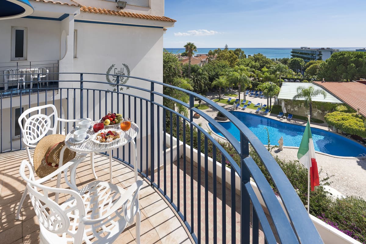 Ein Balkon mit weißen Metallstühlen und einem Tisch, gedeckt mit Obst und einem Getränk, mit Blick auf einen Pool sowie das Meer und Gebäude.