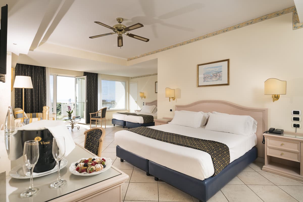 Ein Hotelzimmer mit einem großen Bett, einem Schreibtisch mit einem Laptop, einem Sessel und einem Esstisch mit Kuchen und Weingläsern.