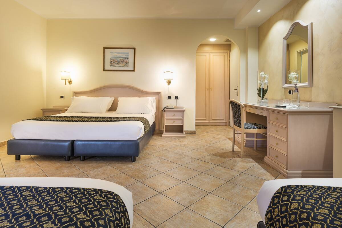 Ein Hotelzimmer mit einem Bett, Nachttischen, einem Schminktisch, einem Spiegel und einer Tür zu einem weiteren Raum.