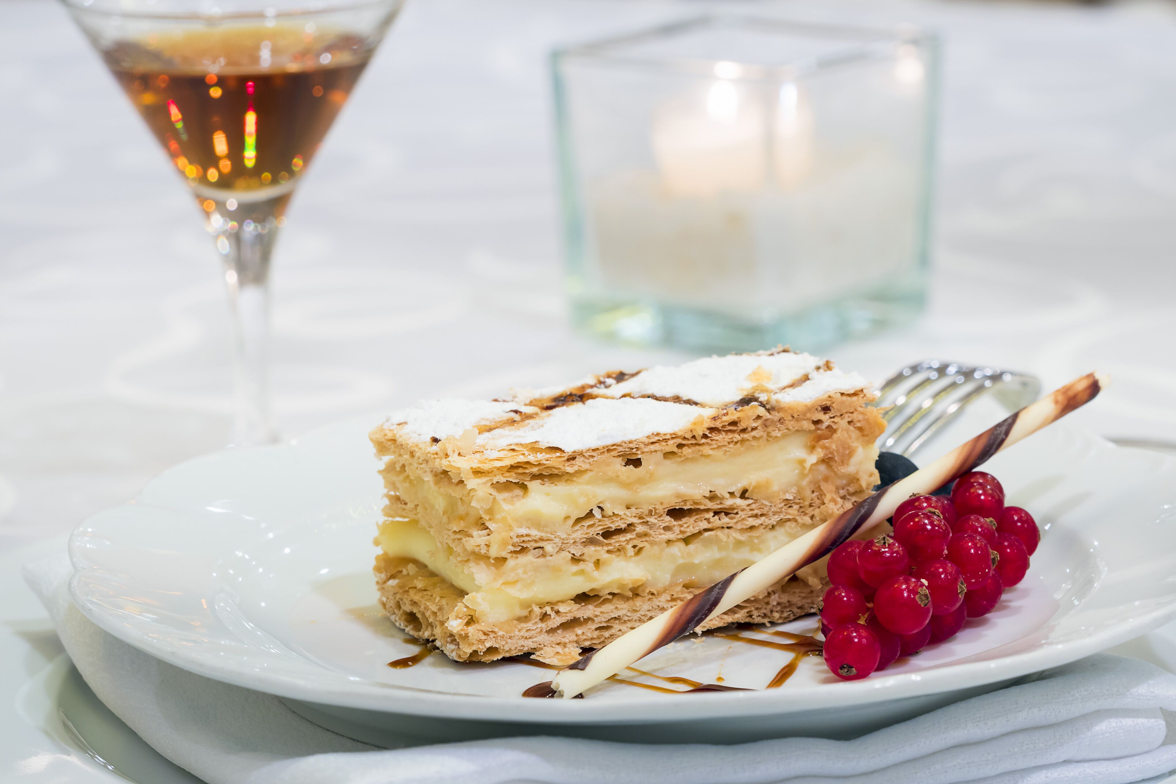 A dessert plate with a layered cake, a glass of a brown beverage, and a candle in the background.