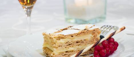 A dessert plate with a layered cake, a glass of a brown beverage, and a candle in the background.