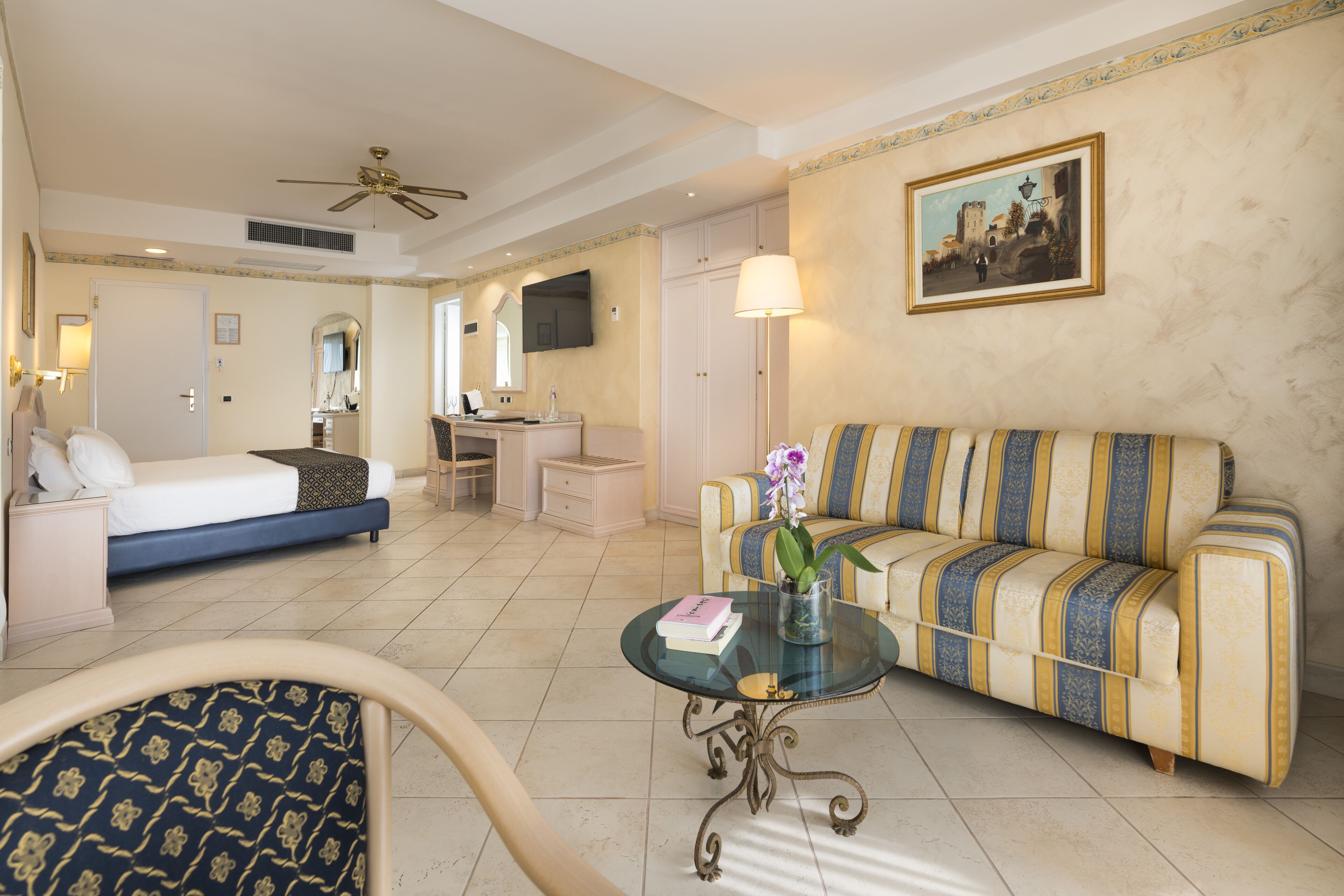 A hotel room with a bed, a sofa, a desk, a chair, a coffee table, and a painting on the wall.