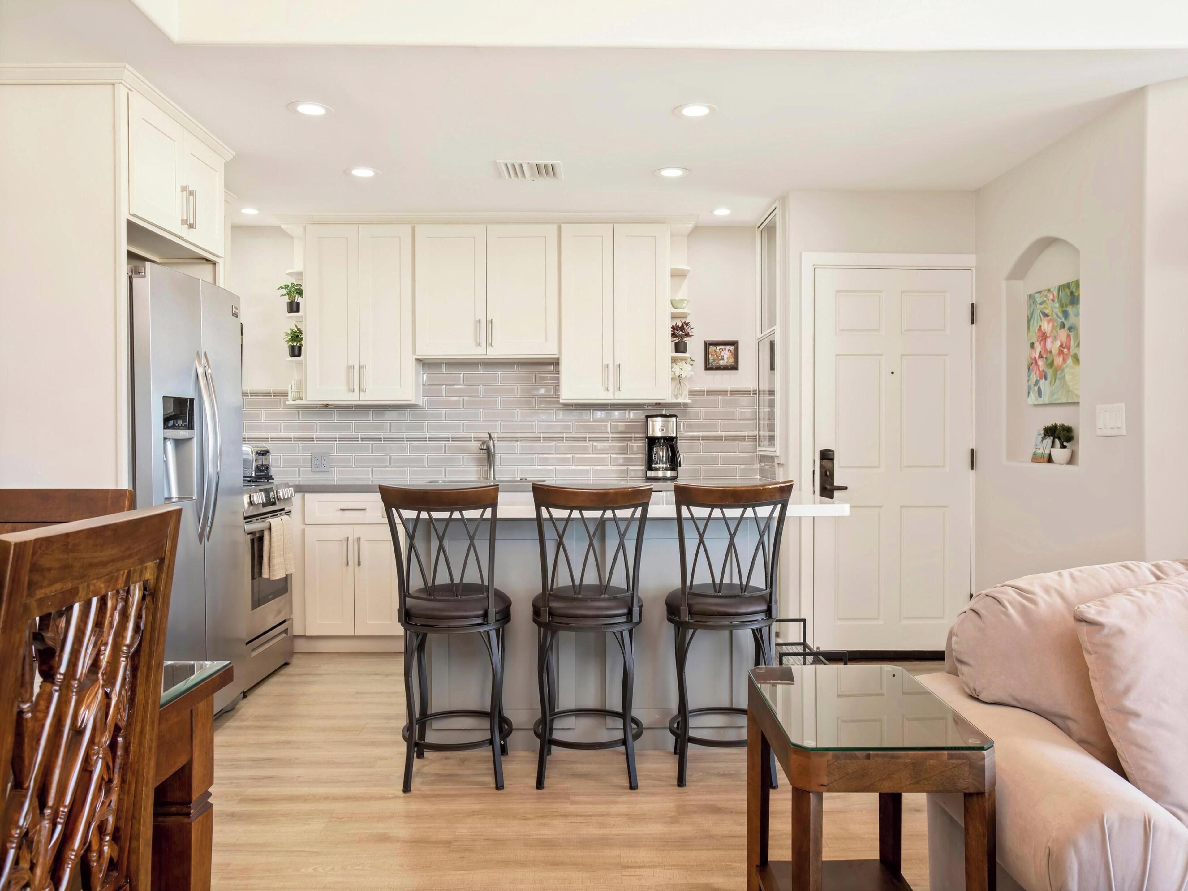 A modern kitchen with white cabinets, a stainless steel refrigerator, and a coffee maker. There are three bar stools and a glass-top table.