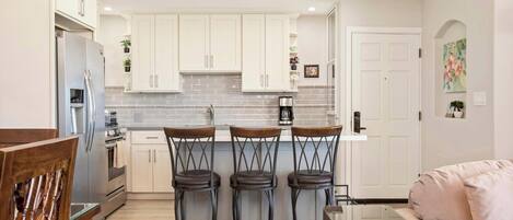 A modern kitchen with white cabinets, a stainless steel refrigerator, and a coffee maker. There are three bar stools and a glass-top table.