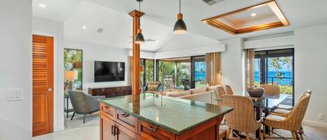 A spacious living area with a large island, wooden cabinets, a flat-screen TV, wicker furniture, and a view of the ocean.