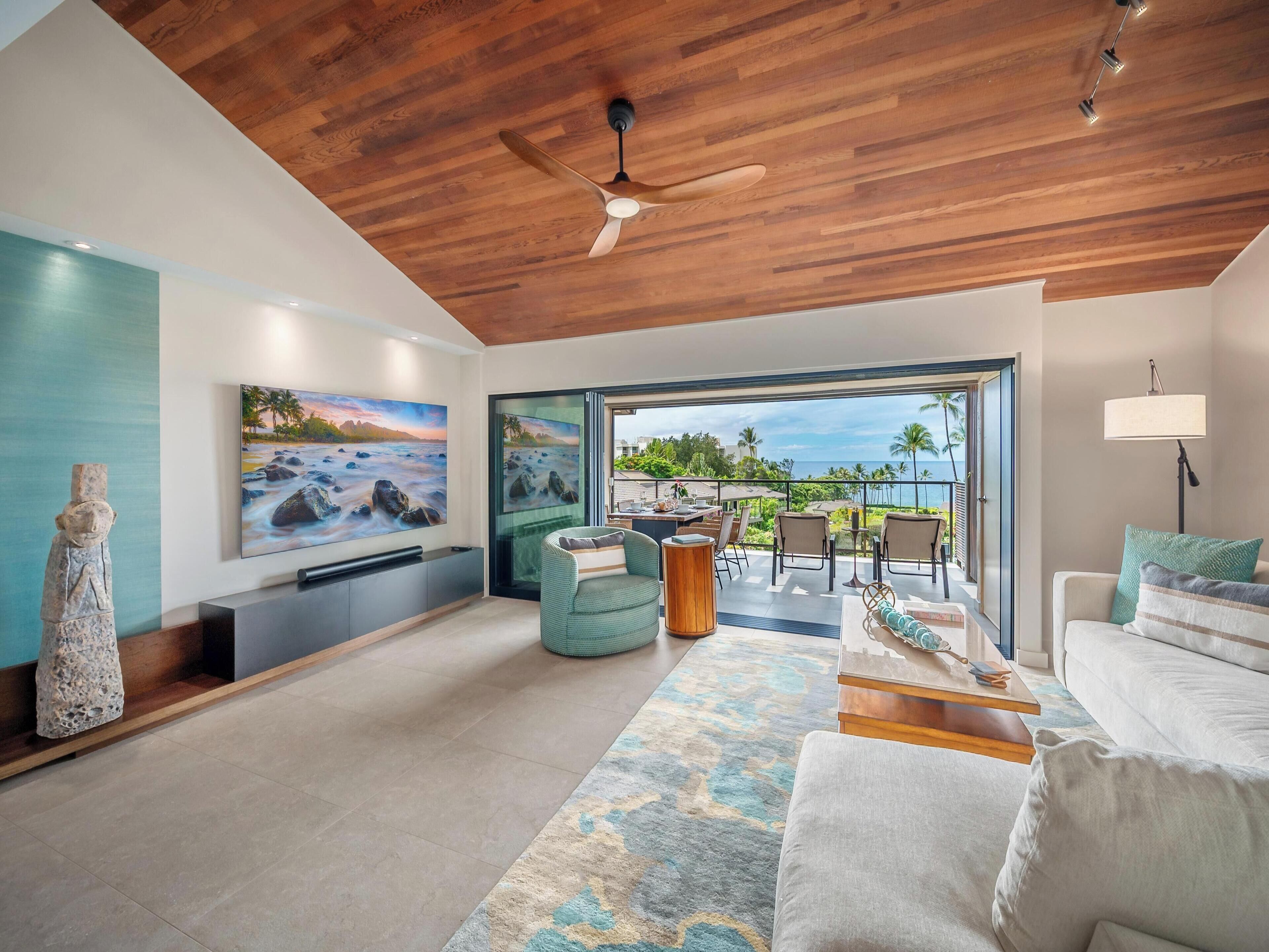 A spacious living area with a wooden ceiling, a large flat-screen TV, a stone sculpture, and a view of the ocean through a glass door.