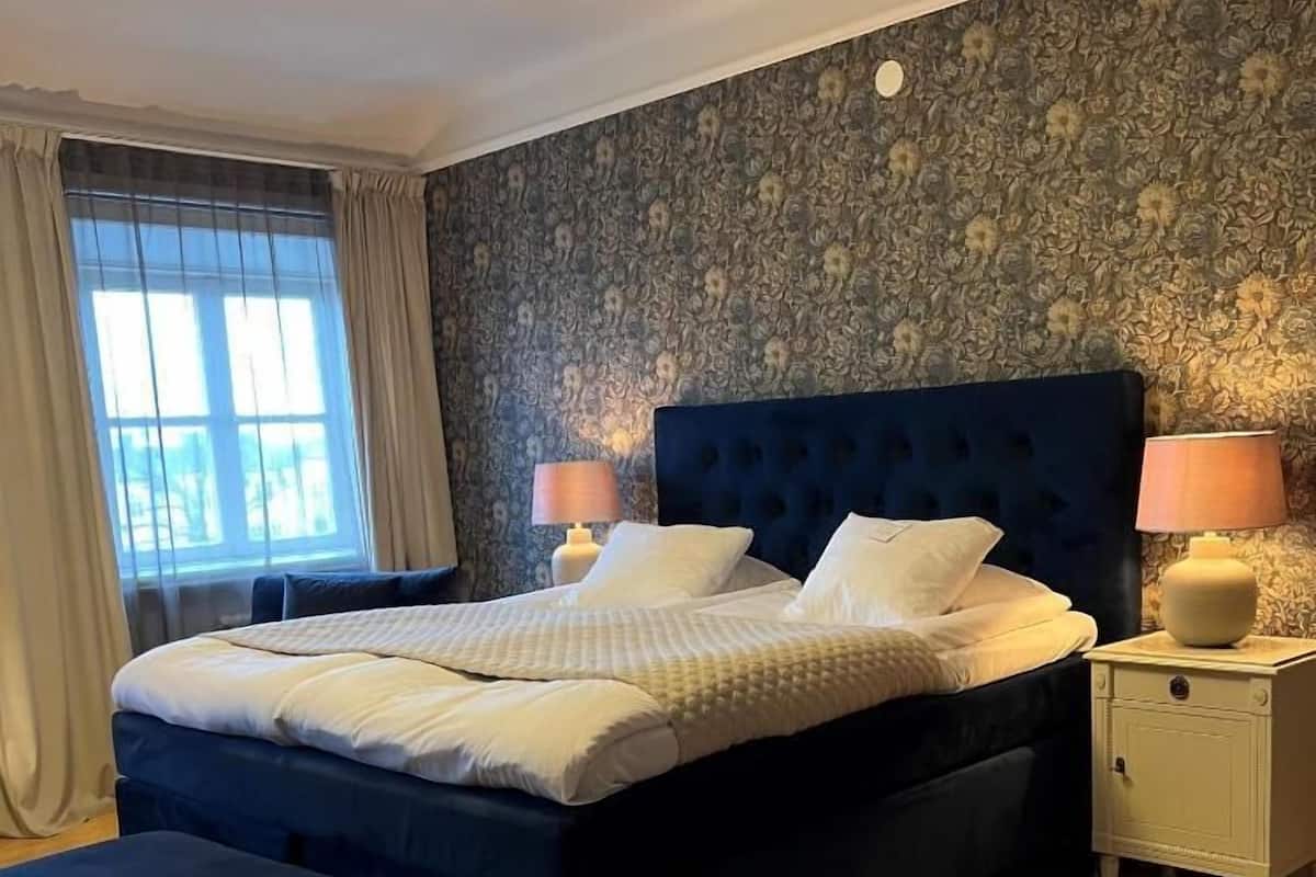 A bedroom with a bed, nightstands, a bench, a window with curtains, and a patterned wall.