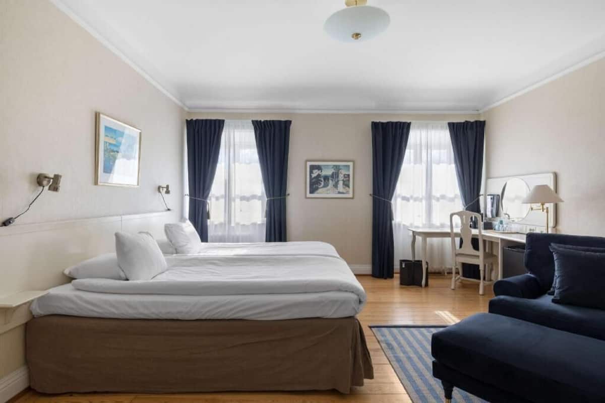 A hotel room with a bed, a desk, a chair, a sofa, and two windows with curtains.