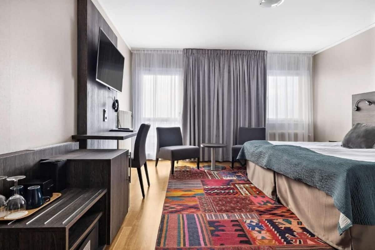 A hotel room with a bed, a desk with a flat-screen TV, two chairs, a small table, and a colorful rug.