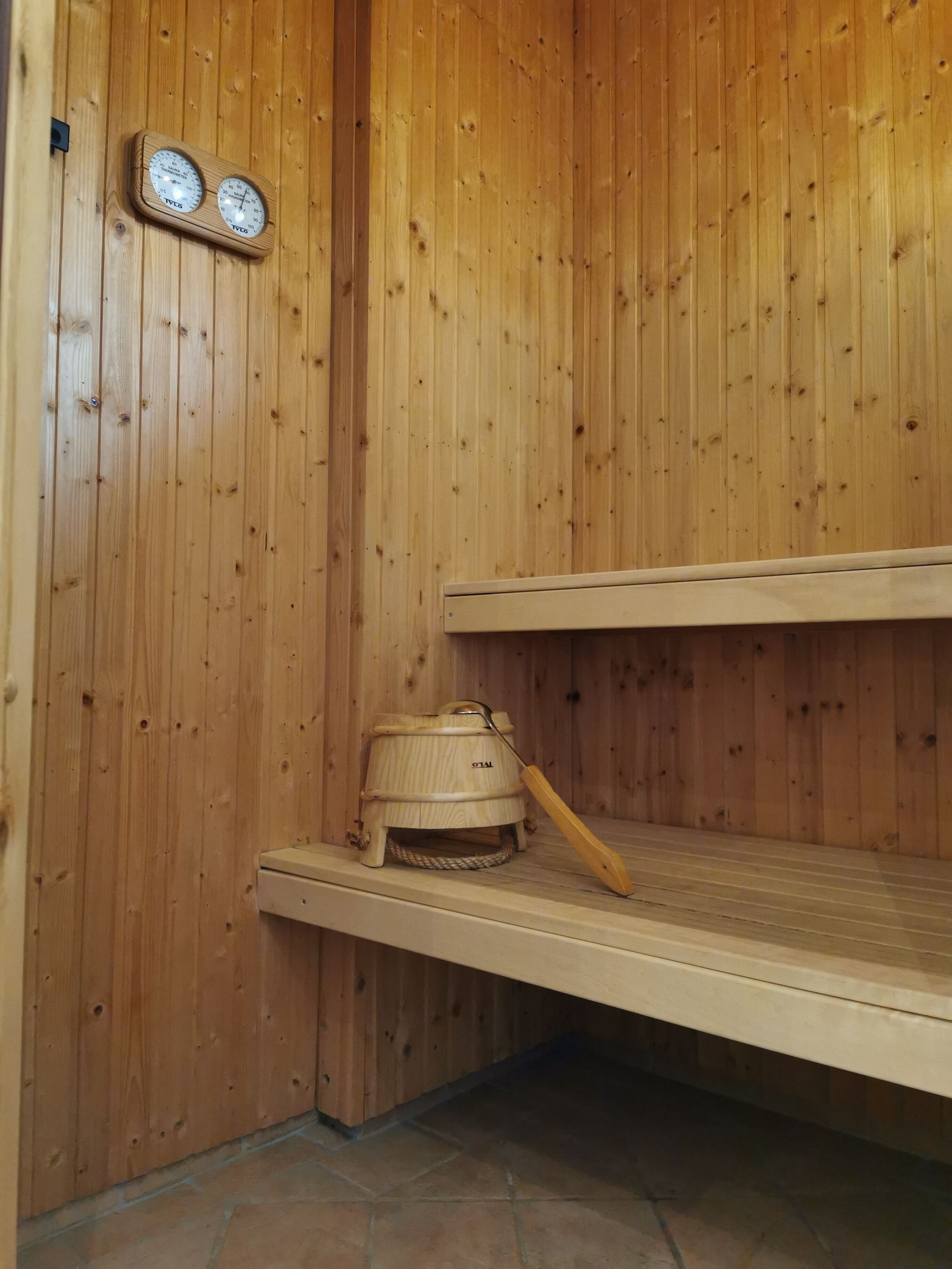 A sauna with wooden walls, a wooden bench, a sauna heater, and two wall-mounted thermometers.
