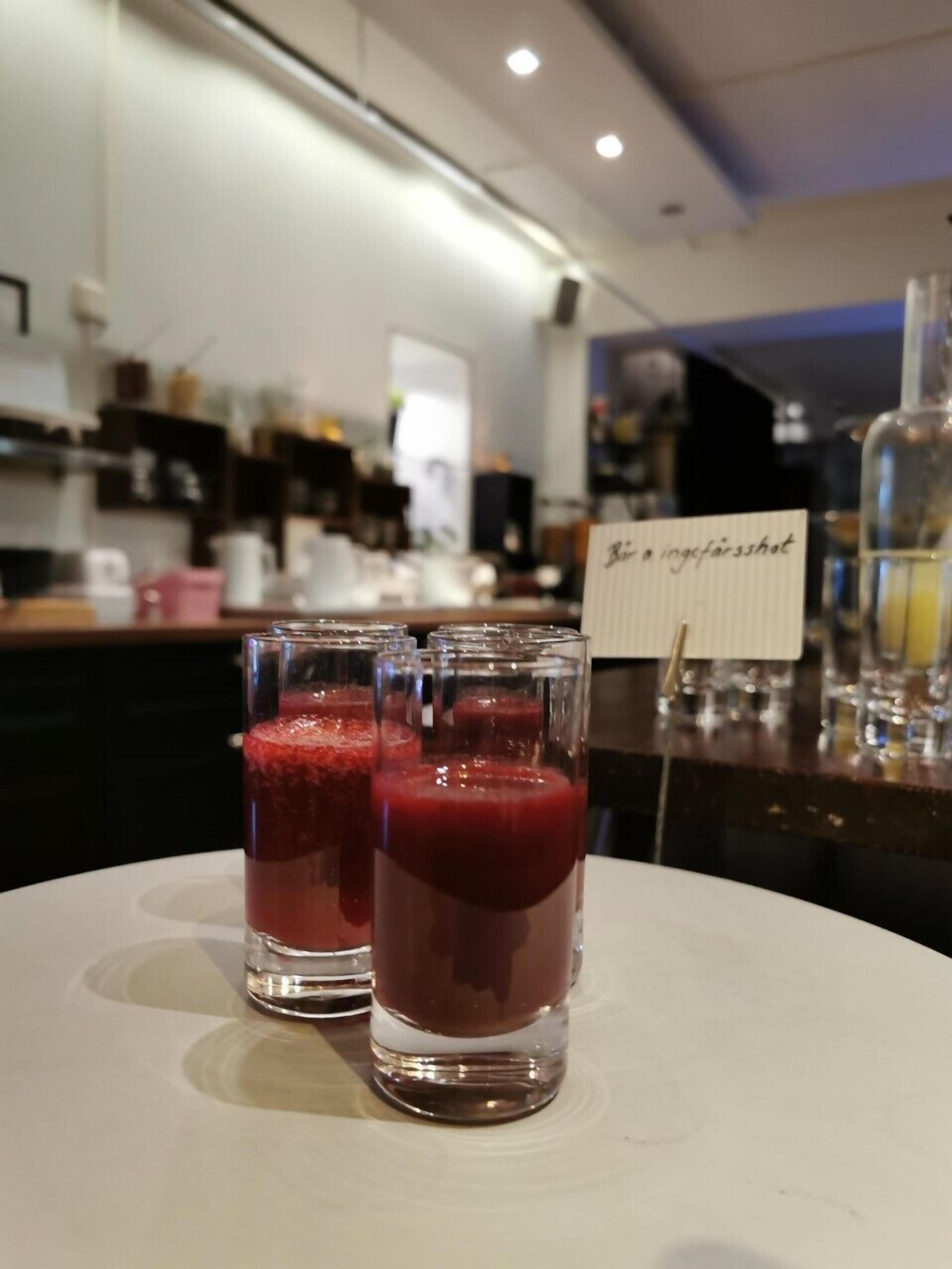 A bar area with a white table displaying two glasses of a red beverage. A sign reads "Bier en ungefarsshot." The background features a kitchen area with various equipment and a counter.