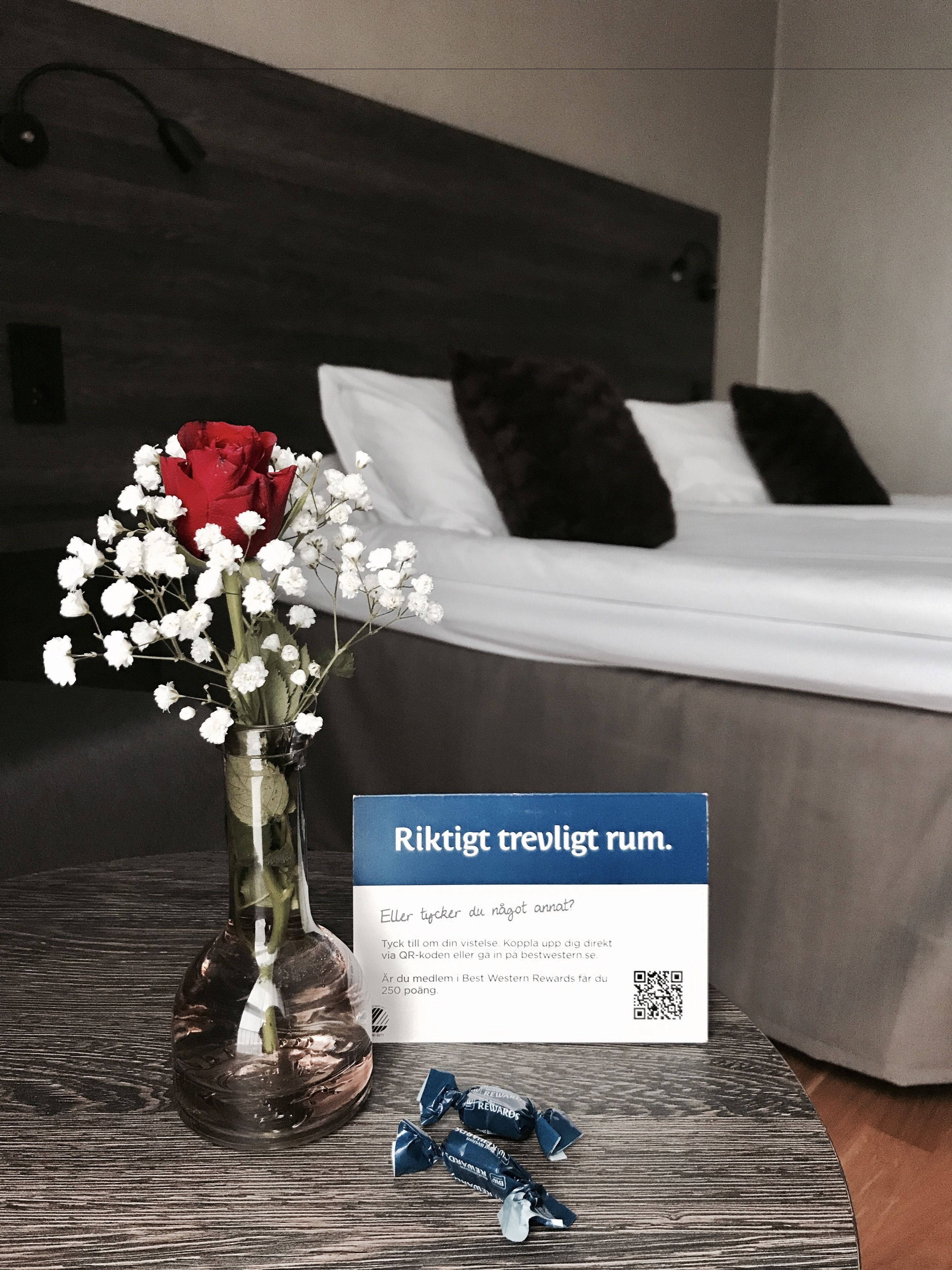 A hotel room with a bed, a bedside table with a vase of flowers, and a sign with a QR code.