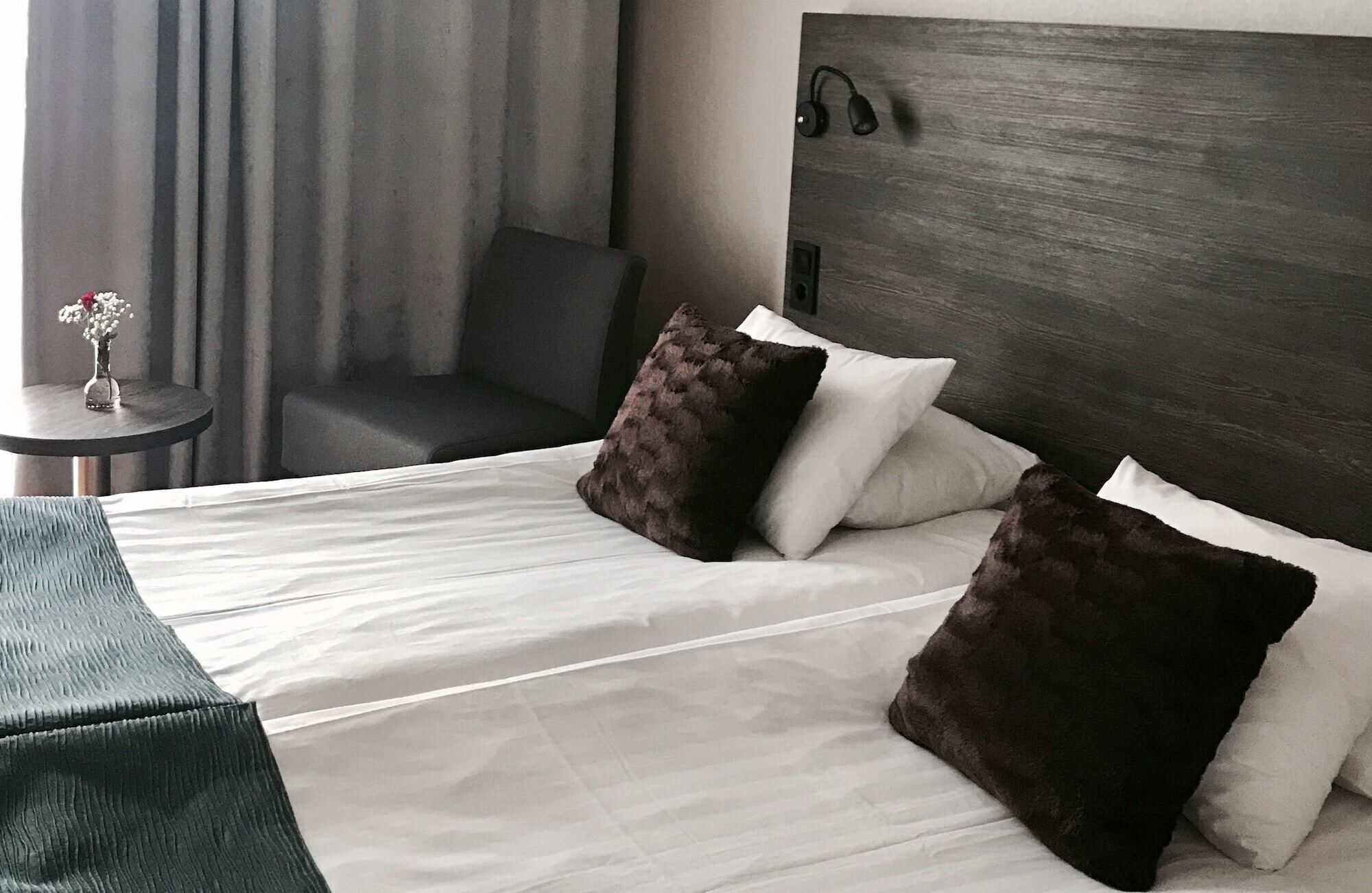 Quarto de hotel com cama, duas almofadas, mesa de cabeceira com um vaso de flores, uma cadeira e cabeceira com alto-falante.