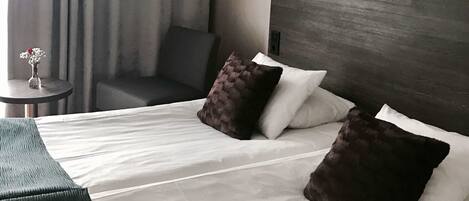 Habitación de hotel con cama, dos almohadas, mesita de noche con un florero y una silla, además de cabecero con altavoz.
