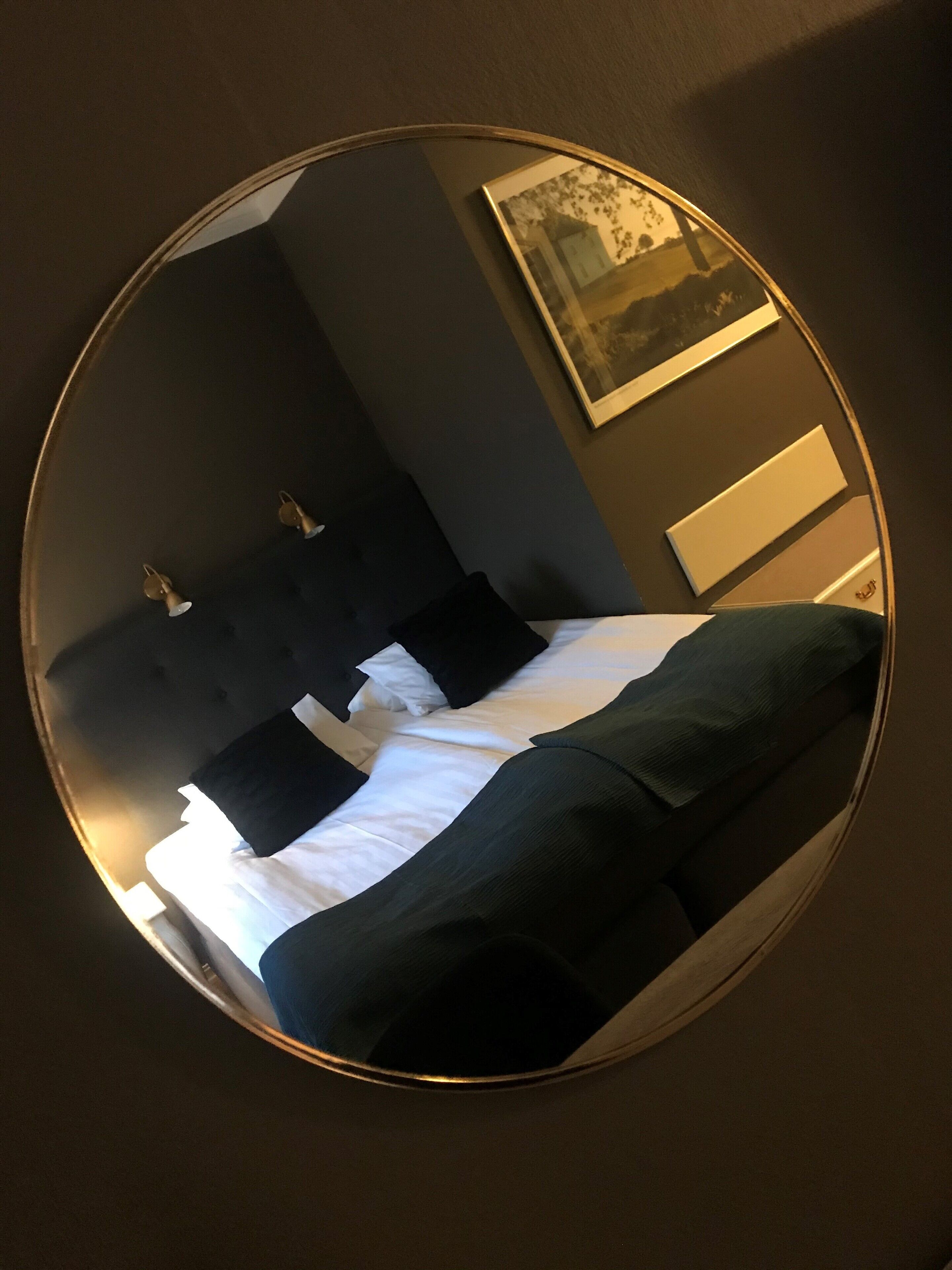 Un espejo circular refleja una cama con cabecera negra y almohadones, una mesita de noche y un cuadro enmarcado en la pared.