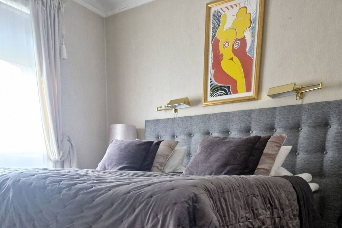 A bedroom with a grey upholstered bed, a grey headboard, a framed artwork above the bed, and a bedside lamp.