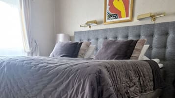 A bedroom with a large bed, grey upholstered headboard, and a framed artwork above the bed.
