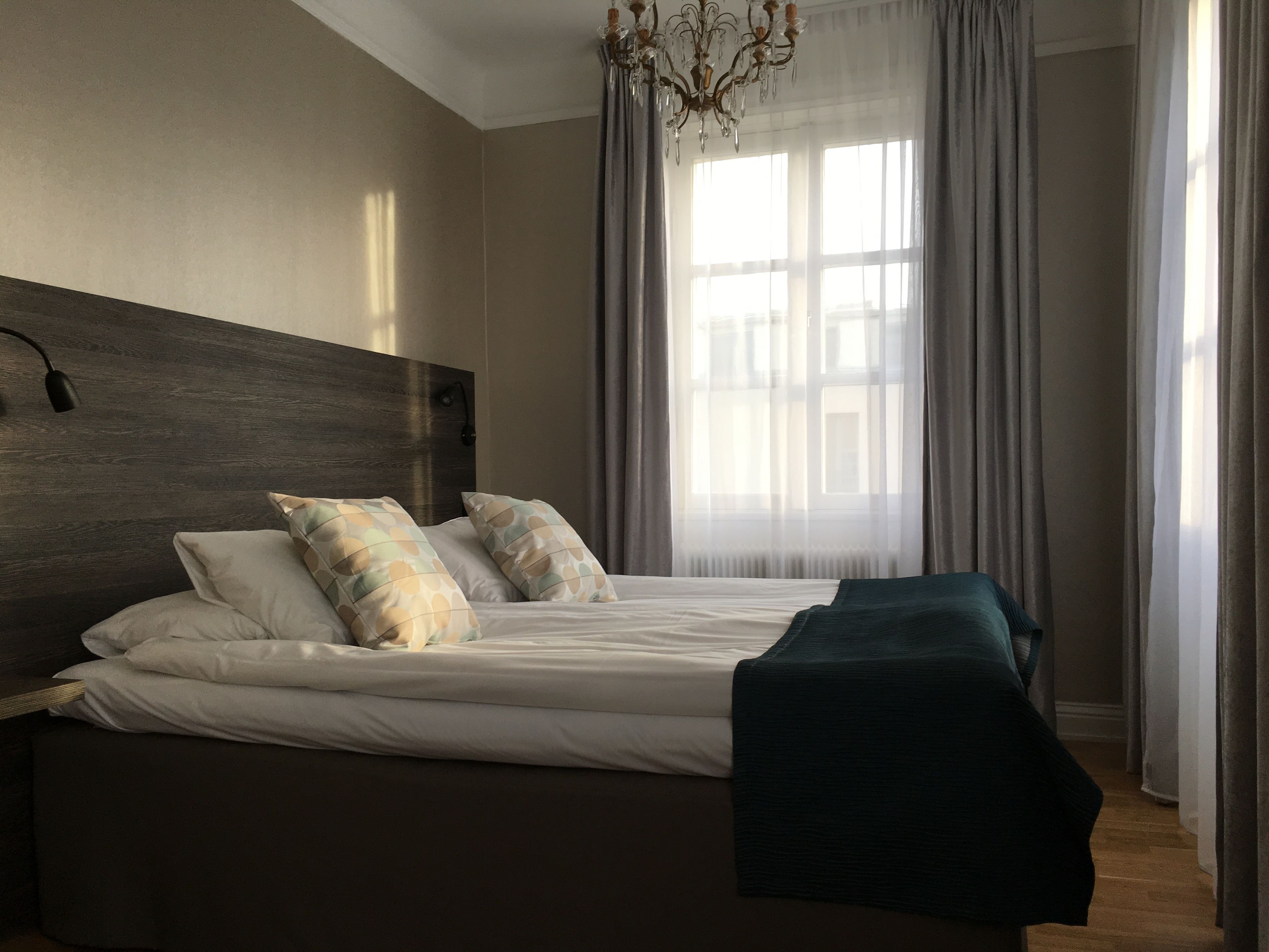 Un dormitorio moderno con una cama grande, cabecera de madera y vistas a un paisaje urbano a través de cortinas translúcidas.