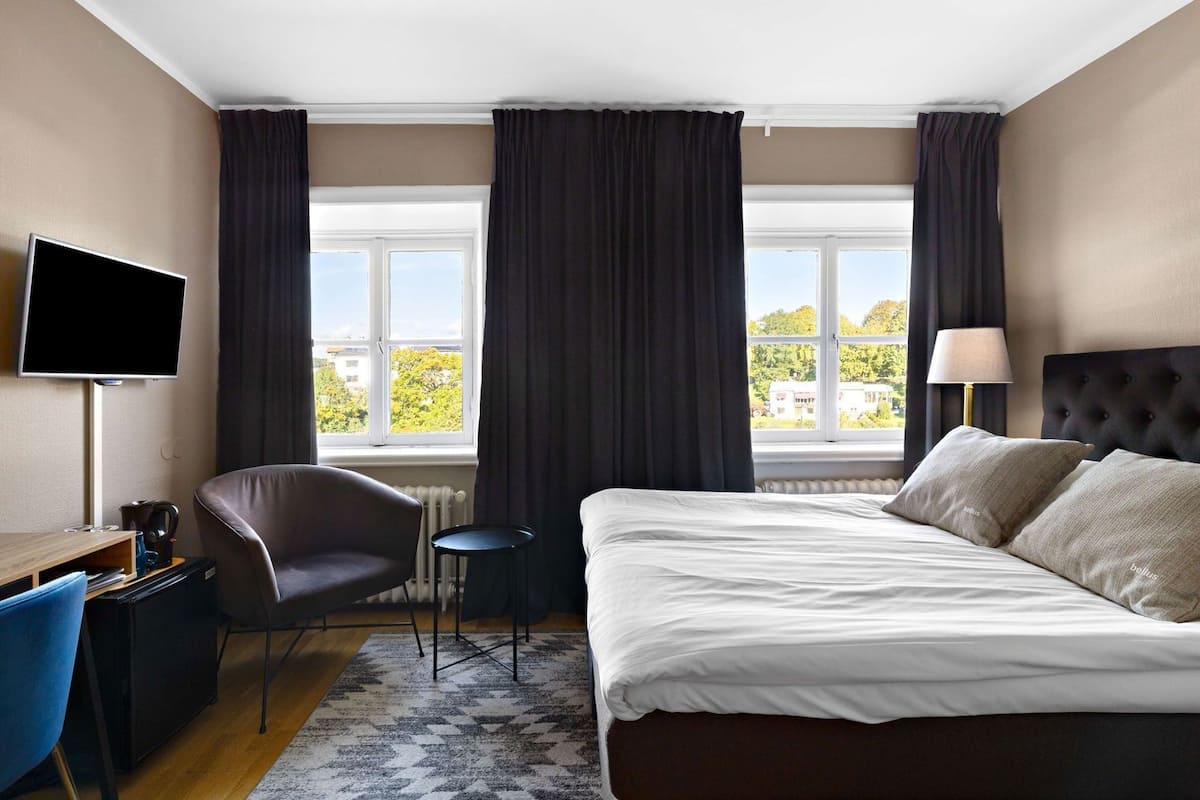 A hotel room with a bed, a desk with a chair, a TV, a lamp, and a view of trees through the window.