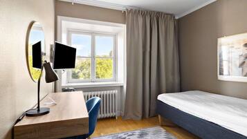 A bedroom with a bed, a desk with a computer monitor, a chair, a window with curtains, and a radiator.