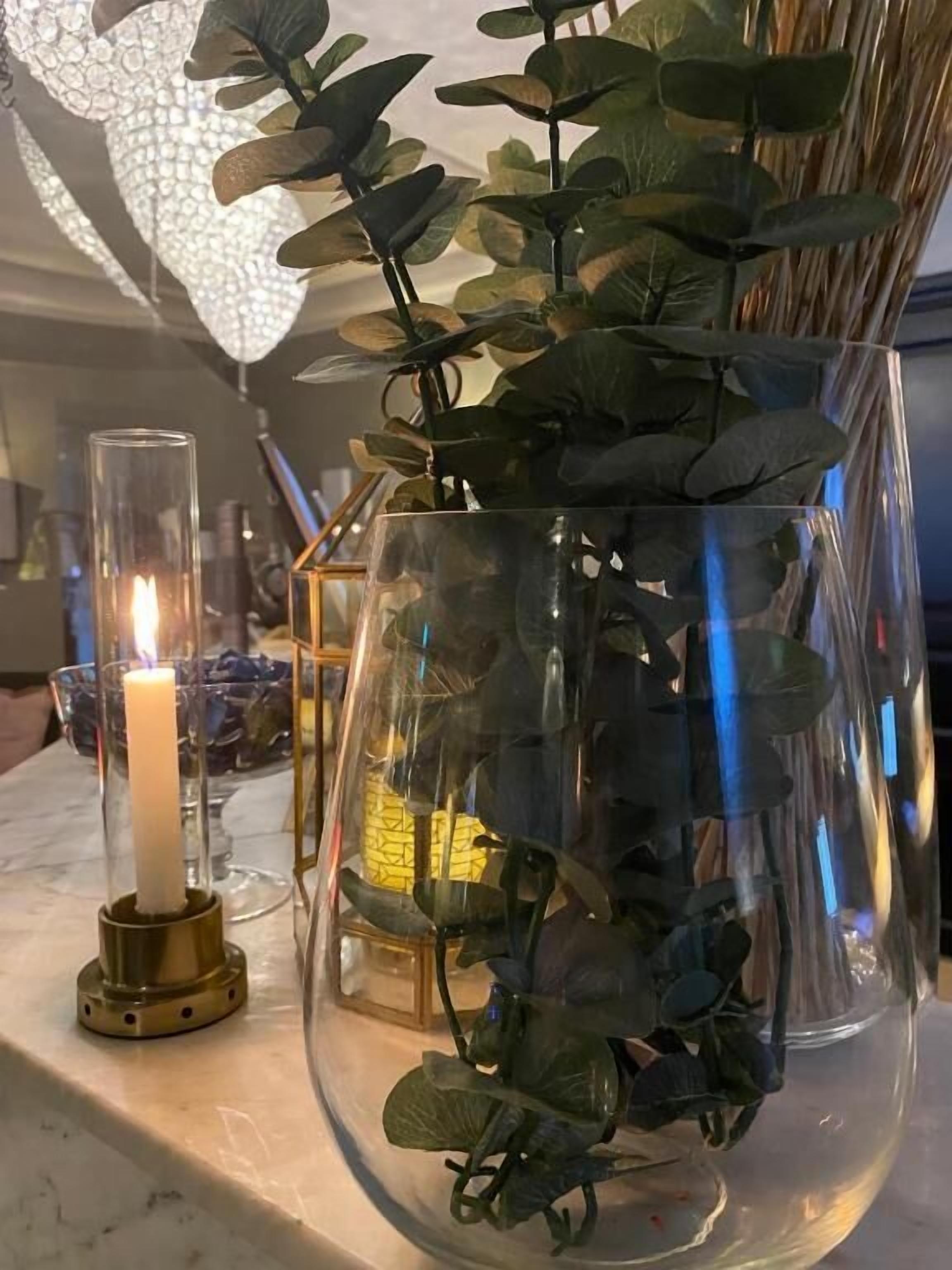 A clear glass vase with green foliage and a lit candle on a marble surface.