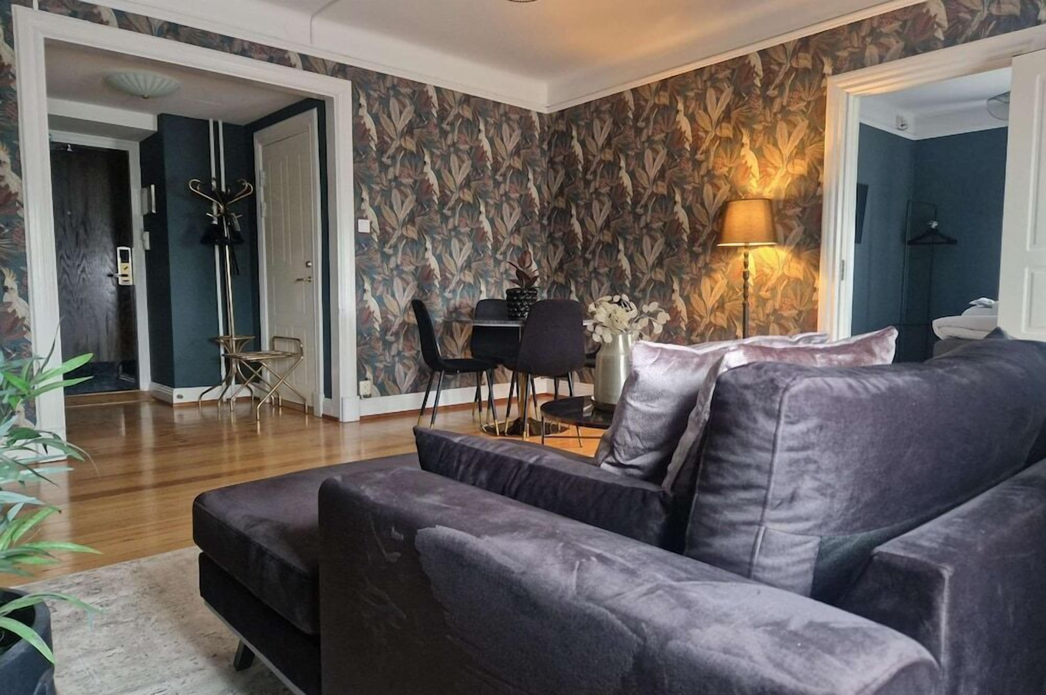 Una sala de estar con un sofá oscuro, una mesa de comedor con sillas y un papel tapiz estampado.