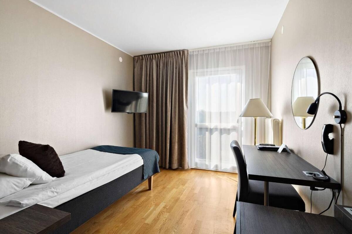 A hotel room with a bed, a desk, a chair, a TV, a lamp, a mirror, and a window with curtains.